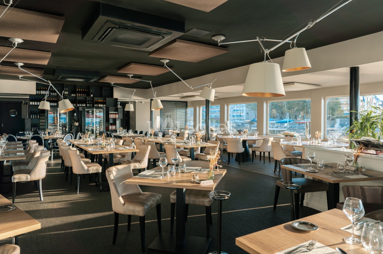 Salle de restaurant intérieur avec tables dressées, chaises beiges, luminaires suspendus et baies vitrées donnant sur la mer.