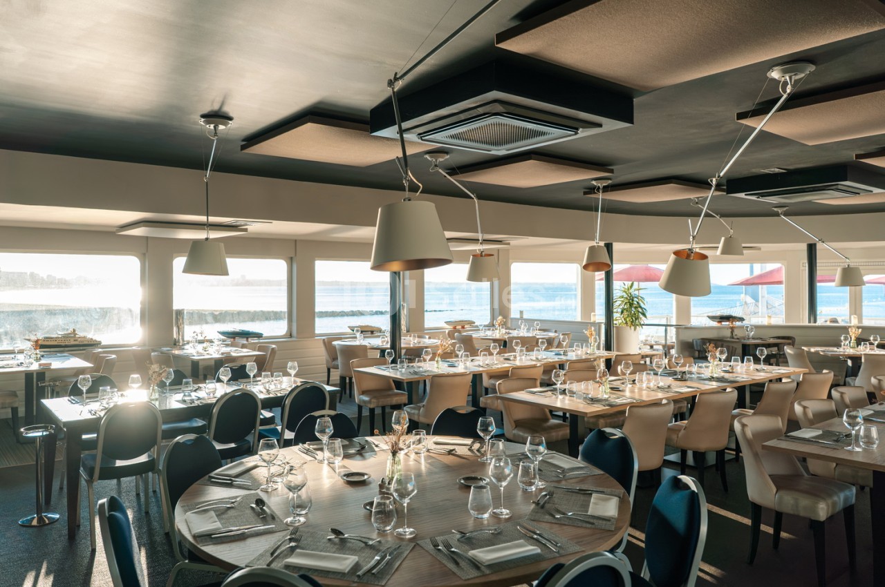 Salle de restaurant dressée avec tables rondes, chaises beige et bleu marine, vue panoramique sur baie et bateaux.
