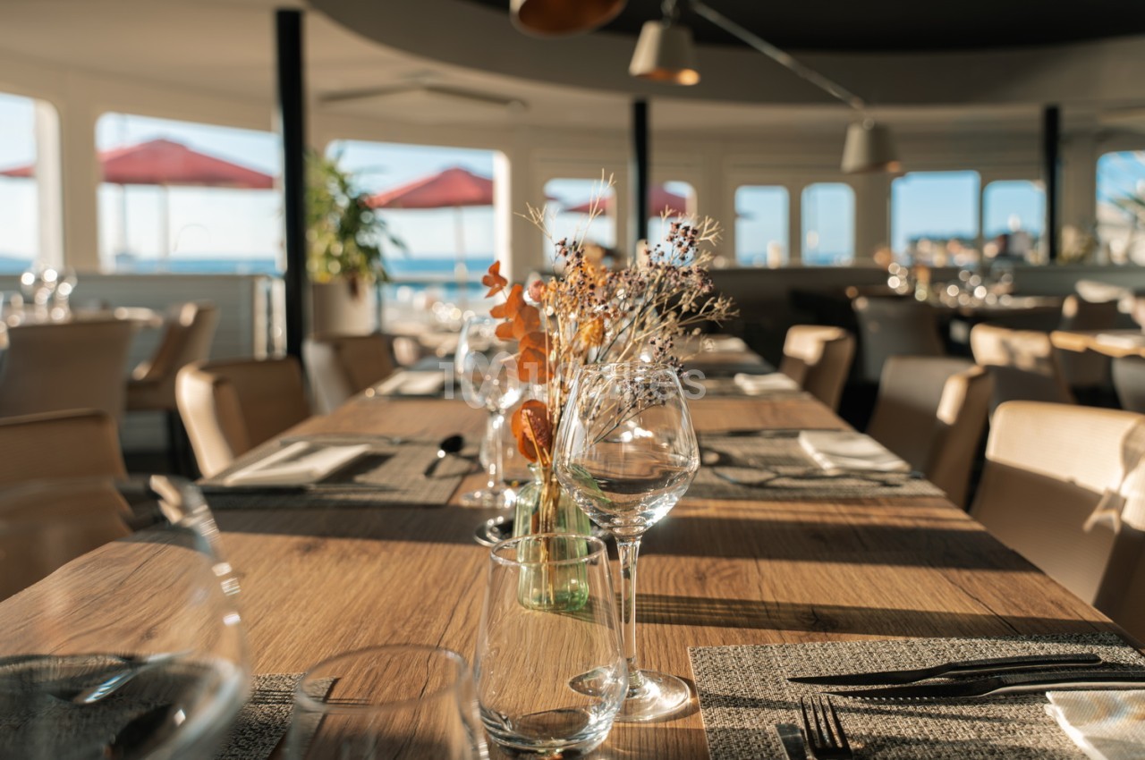 Salle de restaurant avec tables en bois dressées, vue panoramique sur la mer et parasols rouges.