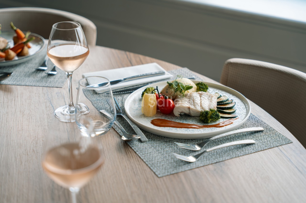 Assiette de poisson blanc garni de légumes et sauce - verre de vin blanc sur table en bois clair.