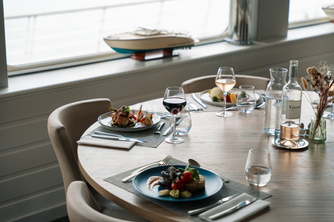 Table dressée avec assiettes bleues et blanches, verres de vin et eau, couverts, vue panoramique sur la mer par les baies vitrées.