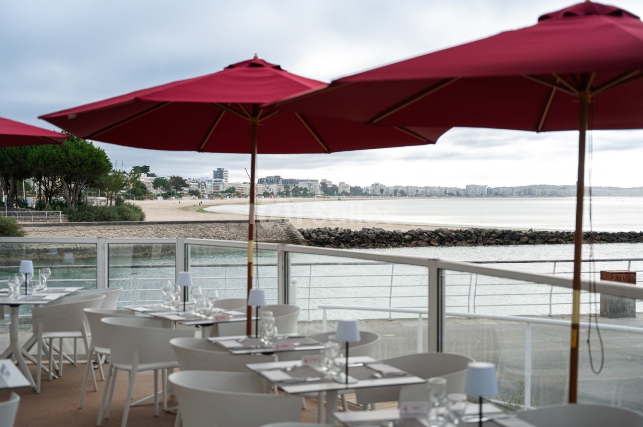 Terrasse de restaurant en bord de plage avec tables dressées sous parasols rouges, vue sur la baie.