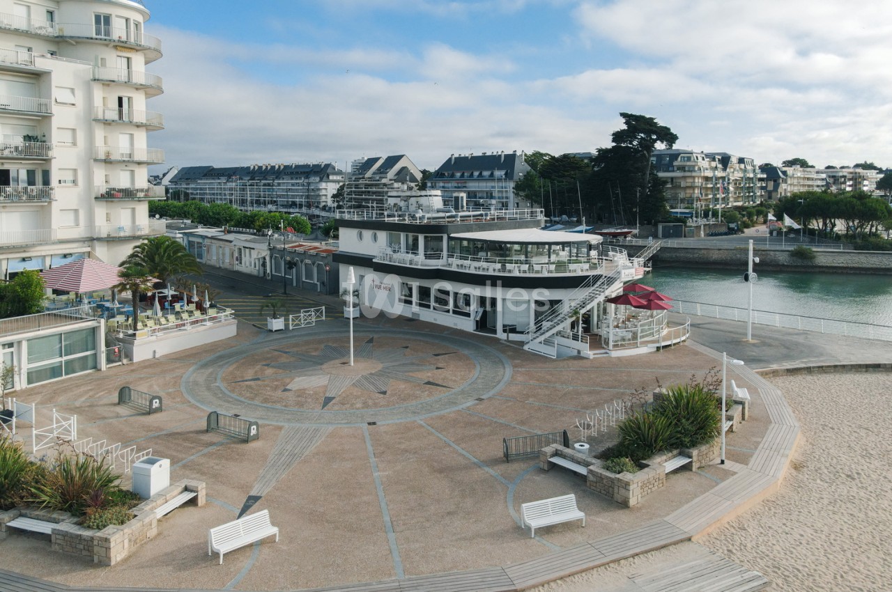 Bâtiment moderne blanc avec terrasse en bord de mer, plage de sable et eau turquoise en arrière-plan.
