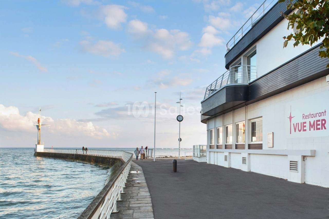 Bâtiment blanc moderne avec terrasse face à la mer et jetée, ciel dégagé.