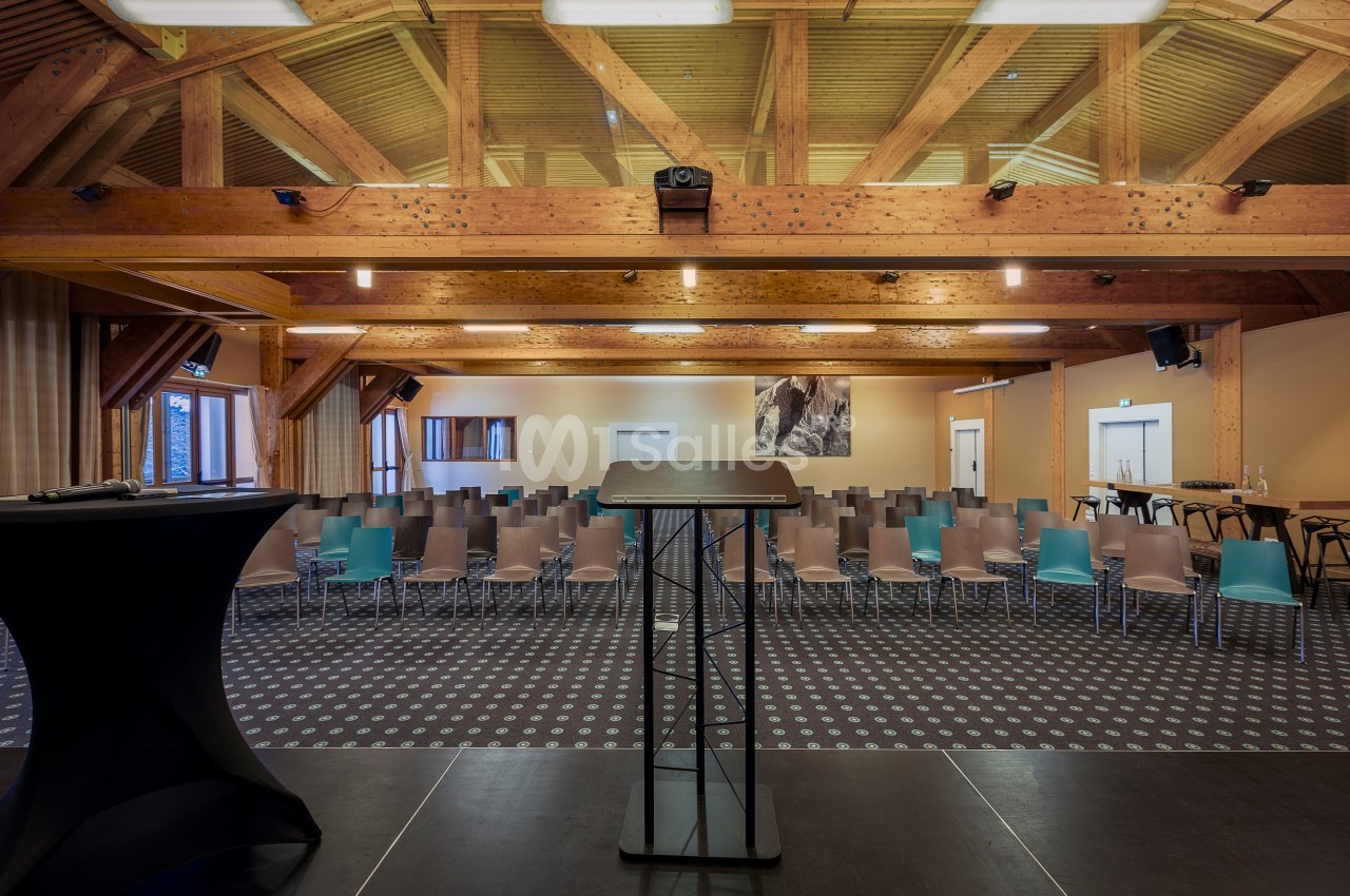Salle de conférence avec estrade, pupitre au premier plan et rangées de chaises vides sous une charpente en bois apparente.