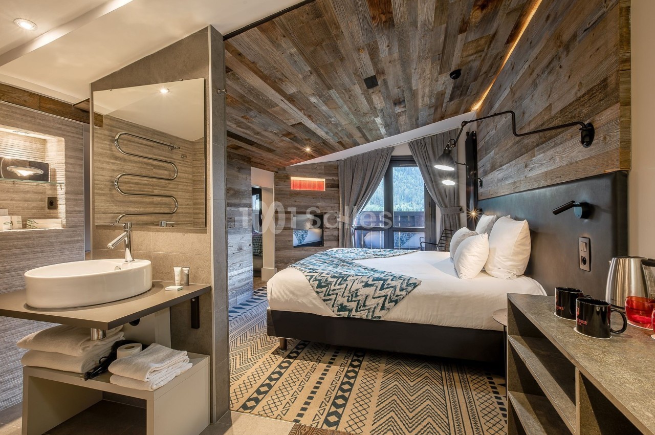 Chambre d'hôtel moderne avec lit double, décor en bois, salle de bain ouverte et vue sur l'extérieur par une baie vitrée.