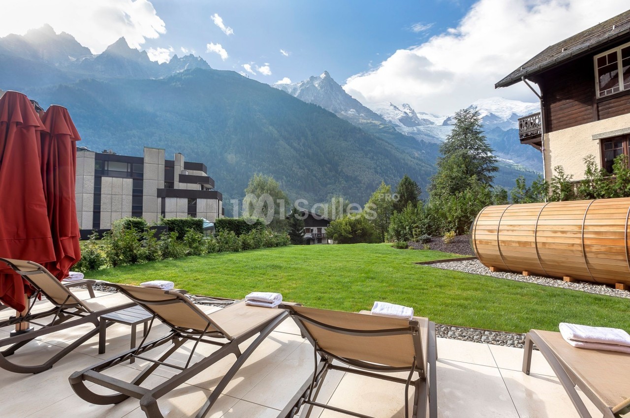 Chaises longues alignées sur une terrasse avec vue sur un jardin, des montagnes enneigées et un sauna en bois.