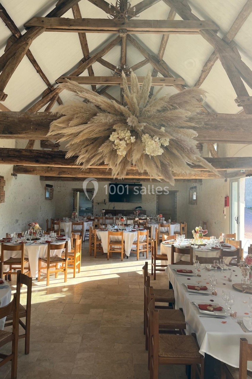 Salle de réception lumineuse avec poutres apparentes, tables dressées et décoration florale suspendue au plafond.