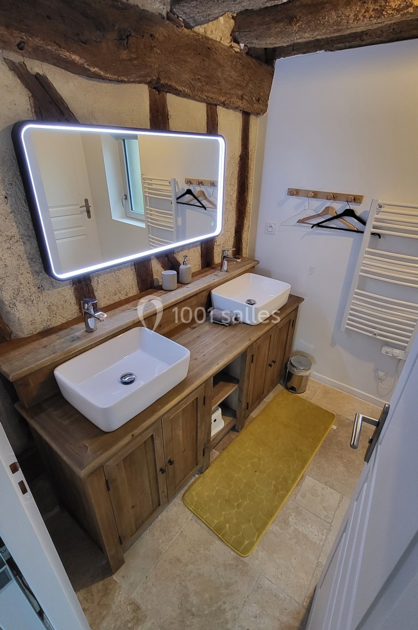 Salle de bain avec double vasque sur meuble en bois, miroir lumineux, radiateur sèche-serviettes et tapis jaune.