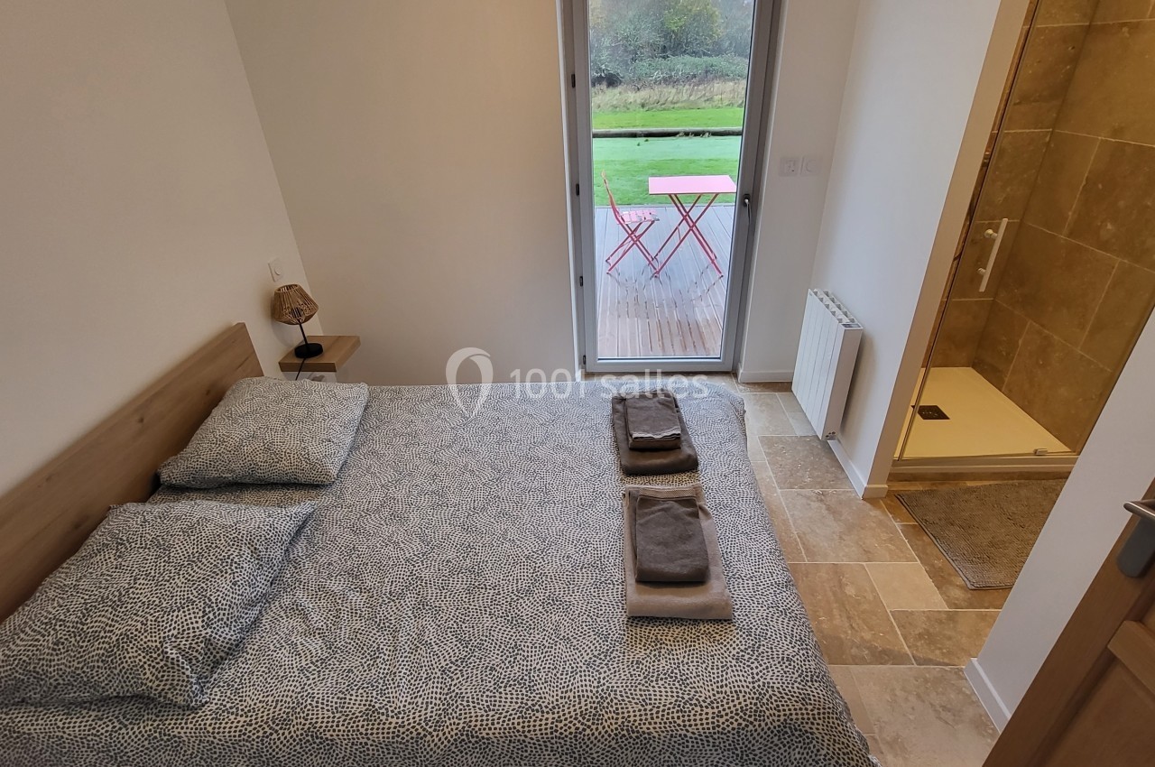 Chambre lumineuse avec lit double, salle d'eau attenante et vue sur une terrasse avec table et chaises rouges.