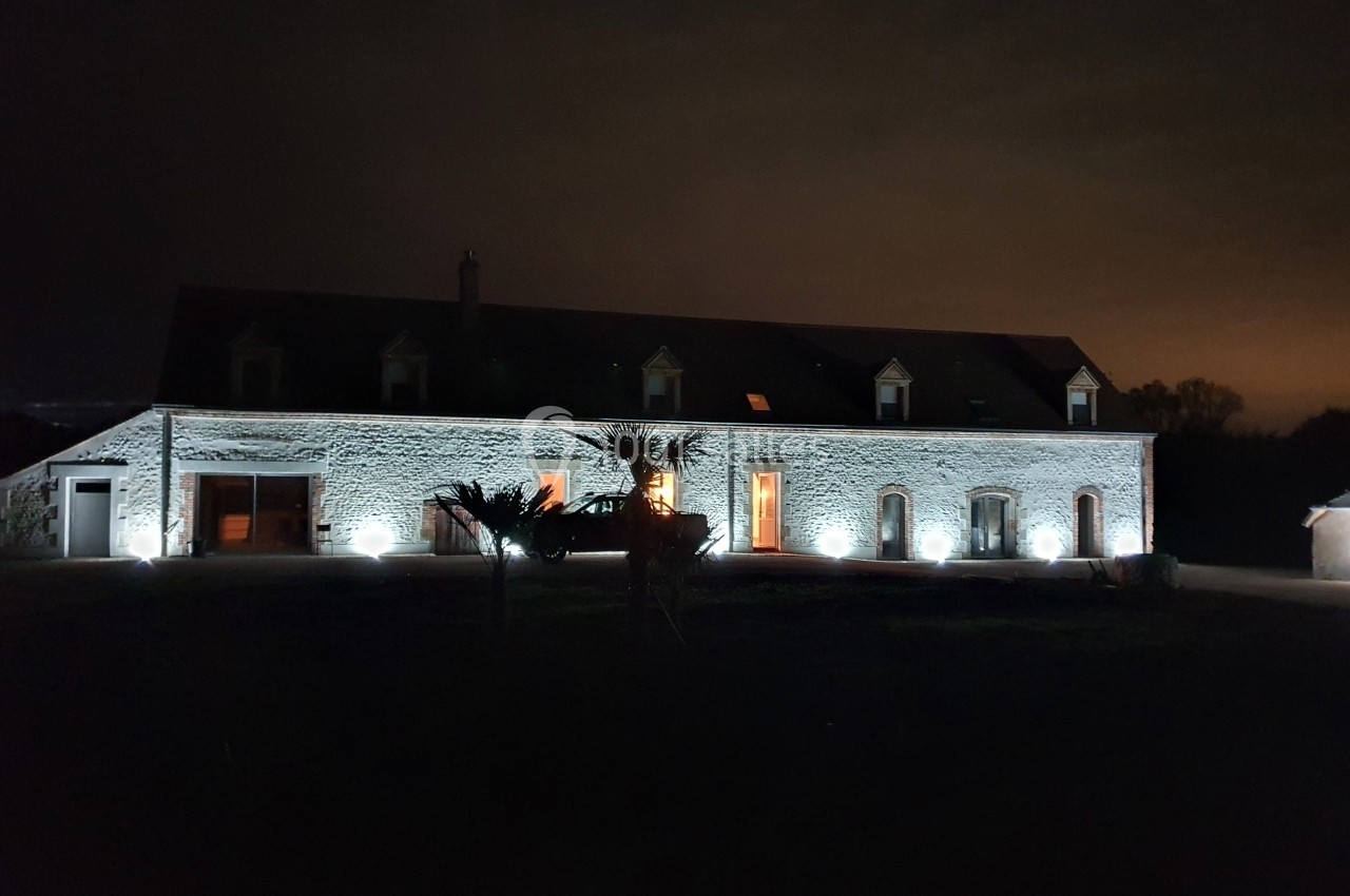 Façade d'une grande maison en pierre éclairée par des lumières extérieures, de nuit, avec un ciel sombre.