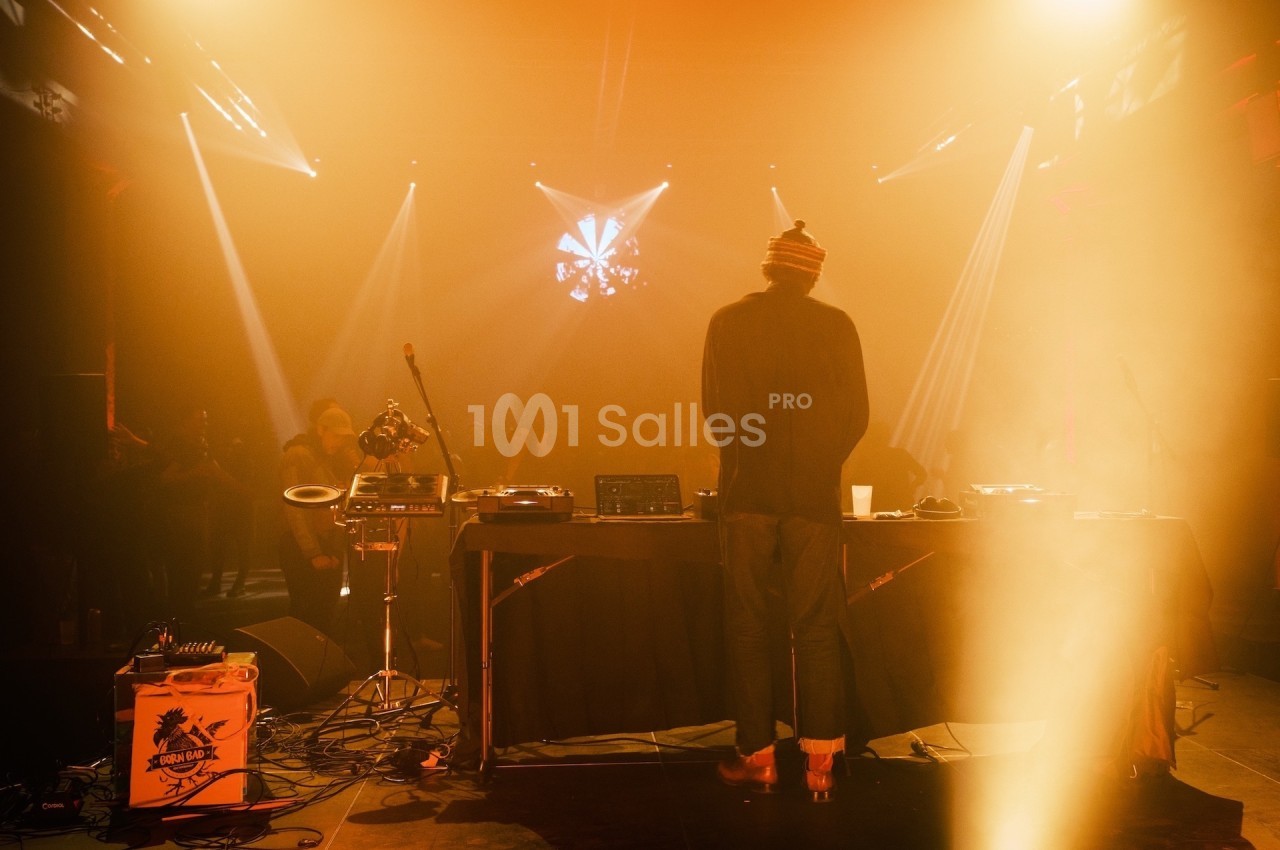 Un DJ de dos mixe sur une scène éclairée par des lumières chaudes, face à un public dans une ambiance tamisée.