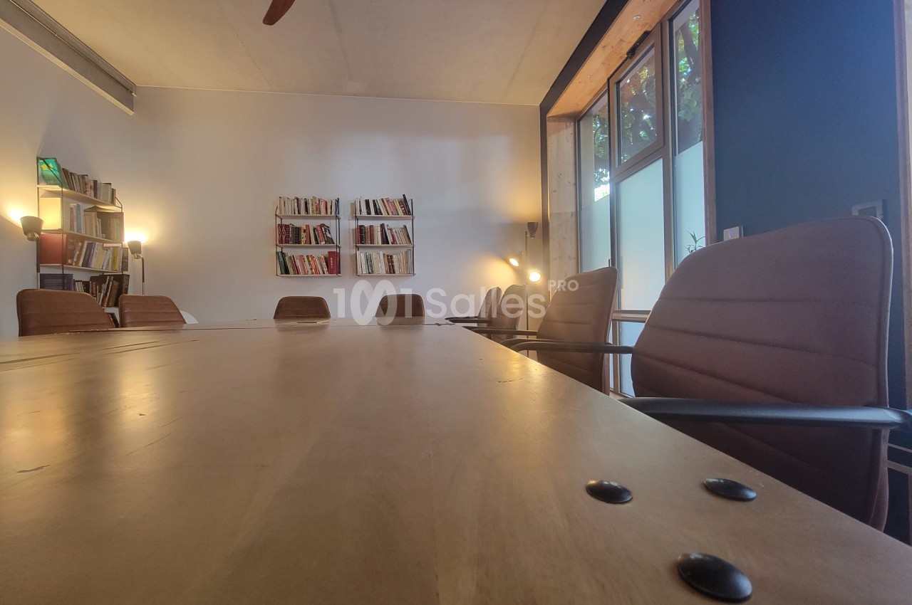 Salle de réunion avec une grande table en bois, chaises en cuir, étagères de livres et lumière naturelle.