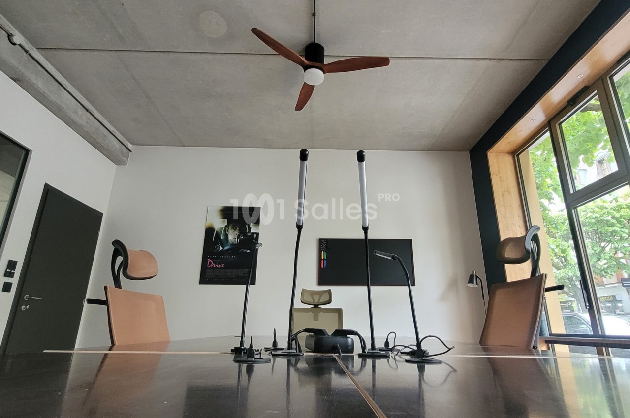 Salle de réunion moderne avec table en bois, chaises design, ventilateur au plafond et grande fenêtre donnant sur des arbres.