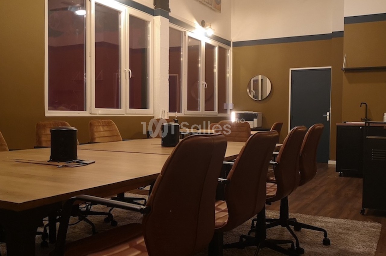 Salle de réunion avec une grande table en bois, chaises en cuir marron, fenêtres et kitchenette en arrière-plan.