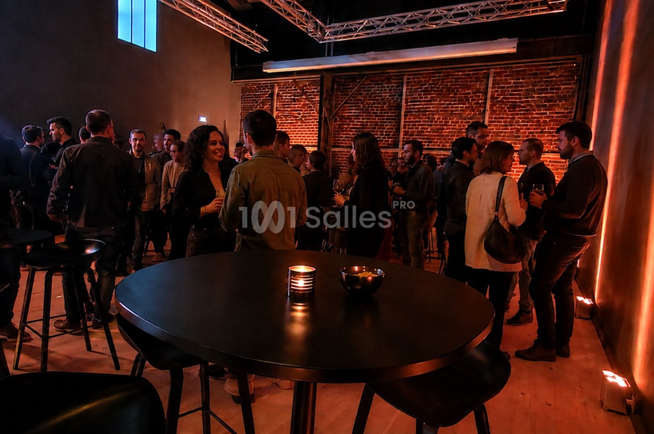 Groupe de personnes discutant dans une salle avec éclairage tamisé, murs en briques et tables hautes.