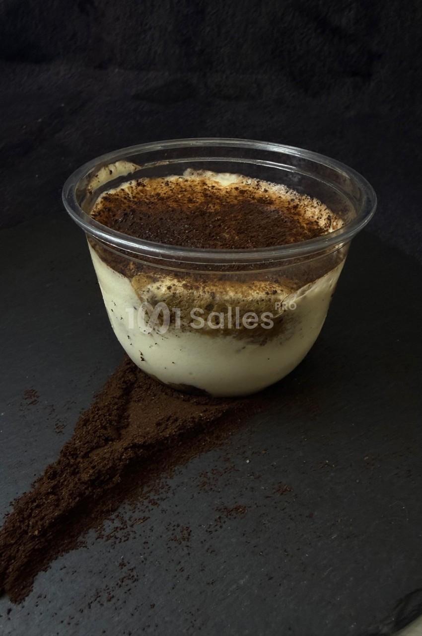Dessert tiramisu dans un pot transparent, posé sur une surface sombre avec de la poudre de cacao répandue.