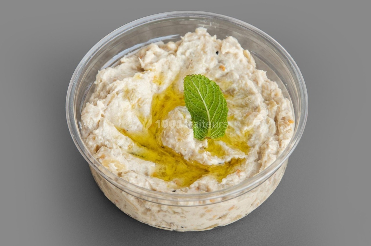 Bol transparent contenant du houmous garni d'huile d'olive et d'une feuille de menthe, sur fond gris.