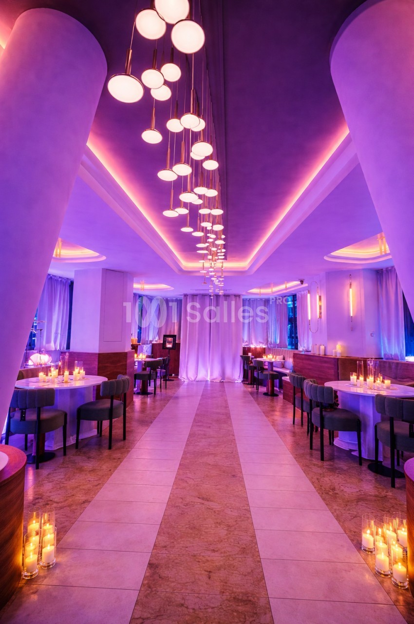 Salle de restaurant élégante avec éclairage violet, tables dressées et bougies disposées au sol.