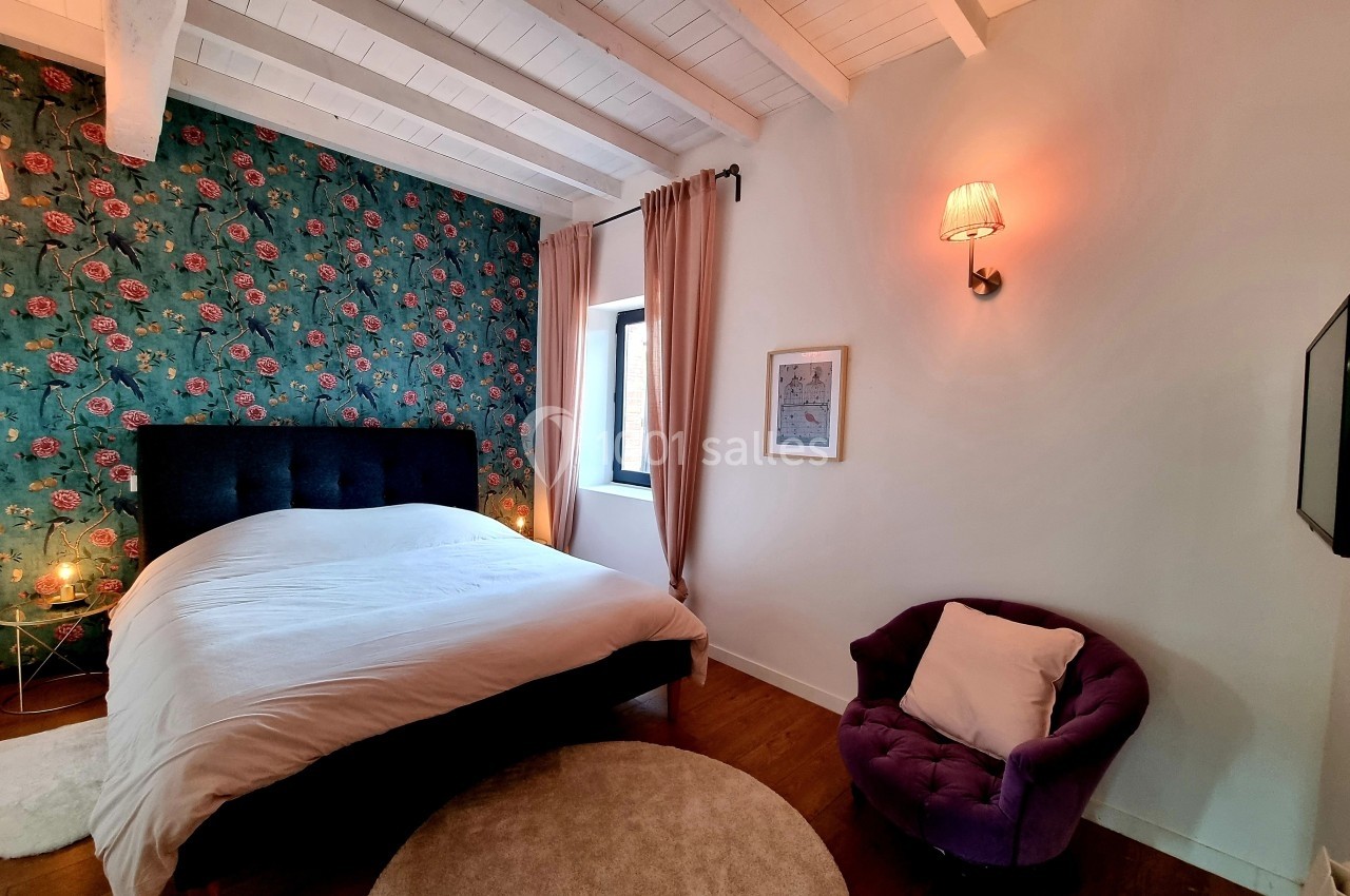 Chambre lumineuse avec lit double, mur fleuri, fauteuil violet, et plafond en bois peint en blanc.