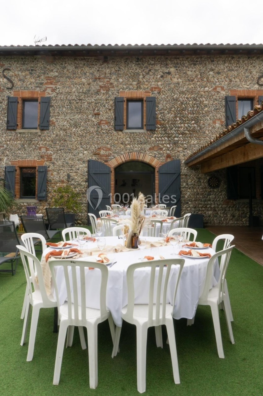 Table dressée en extérieur avec nappes blanches et chaises, devant une maison en pierre avec volets gris.