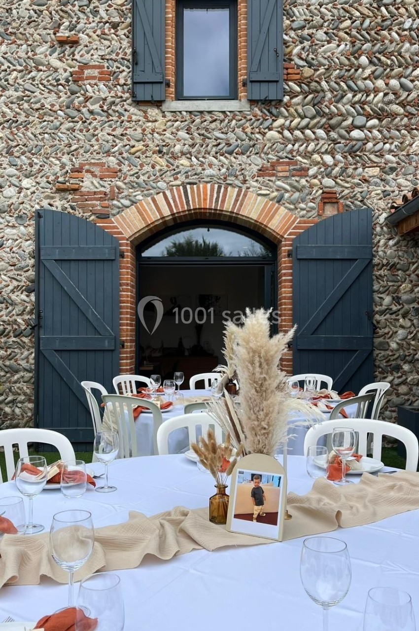 Tables dressées avec nappes beige et décorations florales devant une bâtisse en pierre aux volets bleus ouverts.