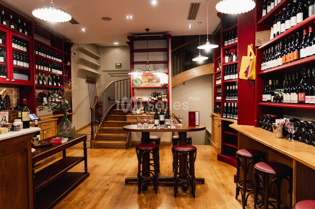 Boutique de vins avec étagères remplies de bouteilles, table centrale avec tabourets et escalier en bois au fond.