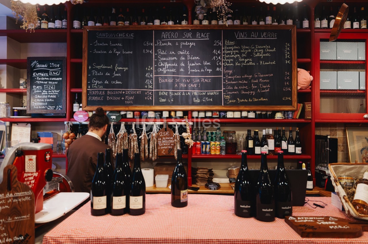 Comptoir d'une épicerie avec bouteilles de vin, charcuterie suspendue et ardoises affichant menus et prix.
