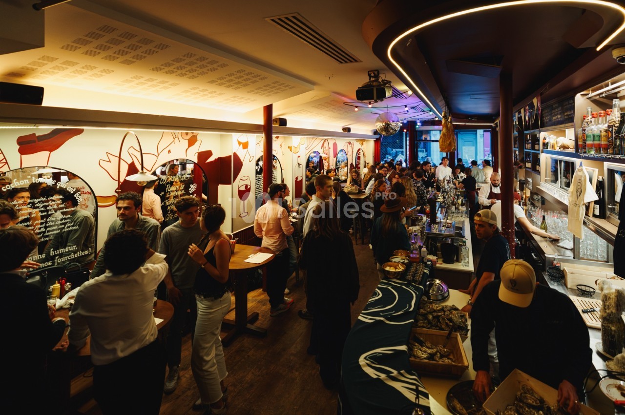 Salle animée d'un bar avec des clients discutant, des décorations murales colorées et un comptoir à huîtres.