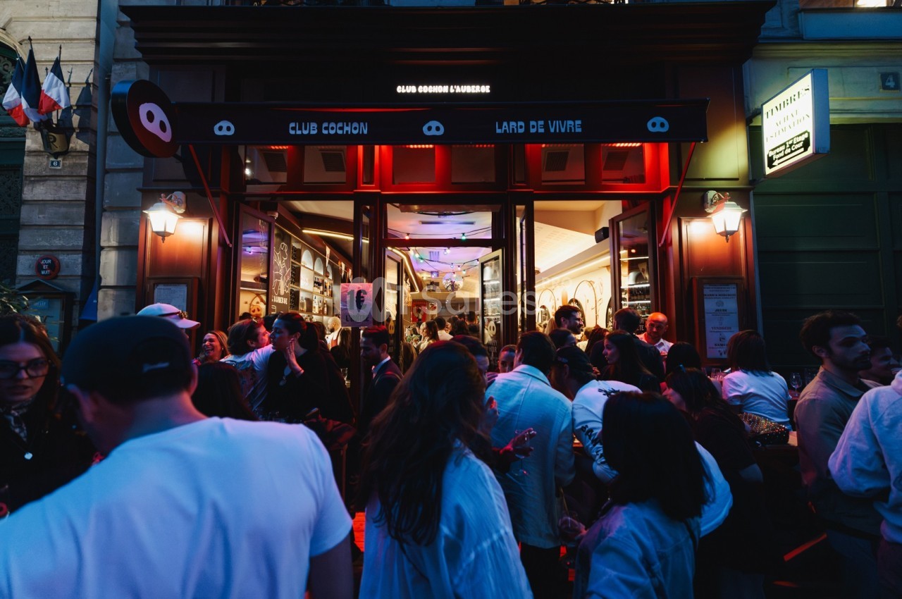 Foule rassemblée devant l'entrée éclairée d'un bar-restaurant en soirée, ambiance animée et conviviale.