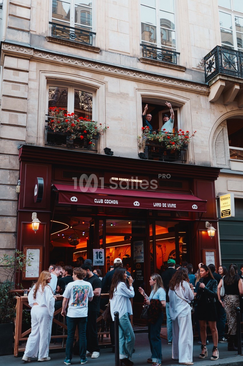 Façade d'un bar animé avec des clients en terrasse et deux personnes saluant depuis une fenêtre au-dessus.