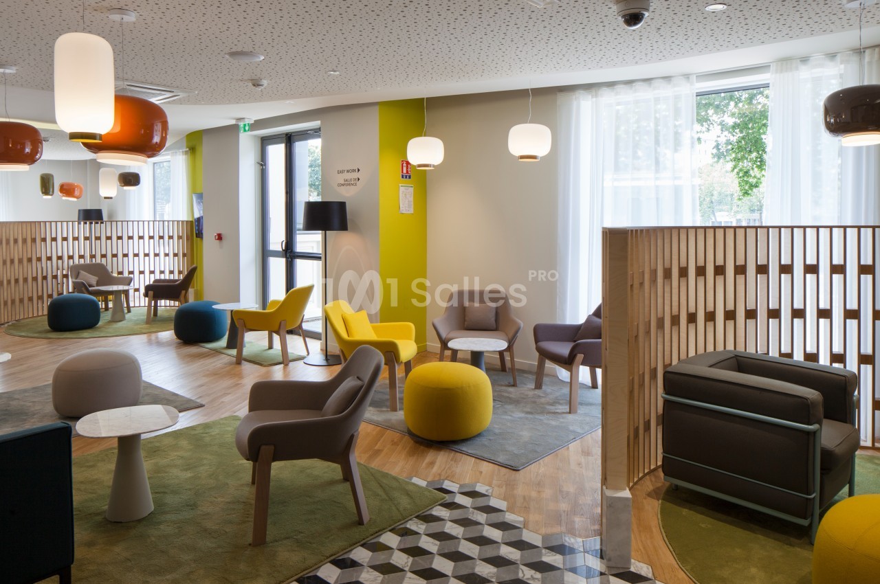 Espace d'attente moderne avec fauteuils colorés, tables basses et lumière naturelle entrant par de grandes fenêtres.