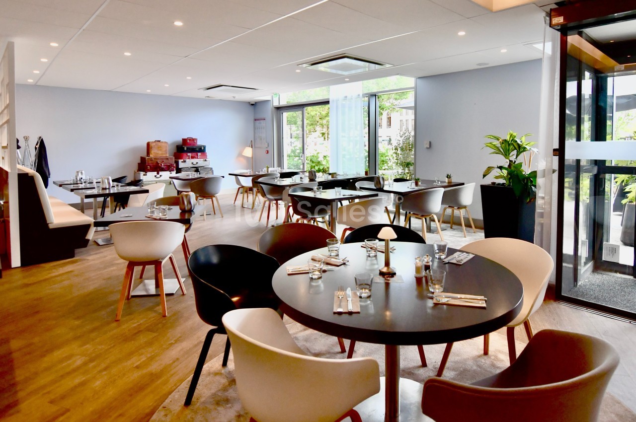 Salle de restaurant lumineuse avec tables dressées, chaises modernes et grandes fenêtres donnant sur un espace vert.