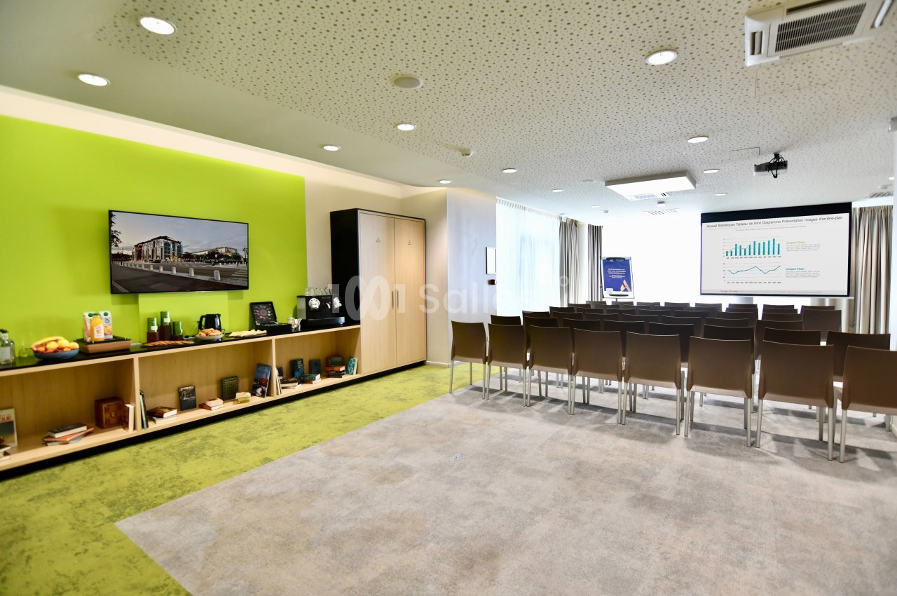 Salle de conférence lumineuse avec chaises alignées, écran de présentation, étagères et mur vert en arrière-plan.