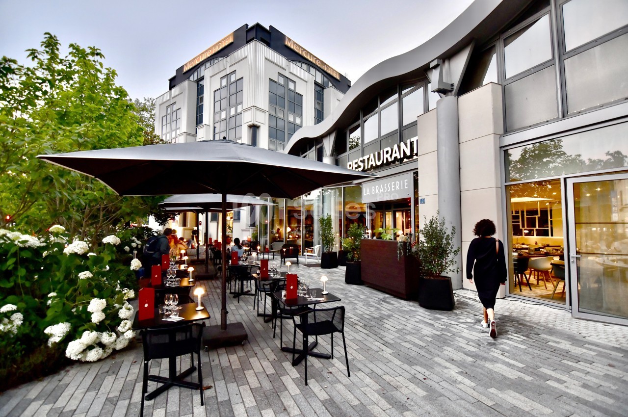 Terrasse d'un restaurant avec tables dressées, parasols et végétation, située devant un bâtiment moderne.