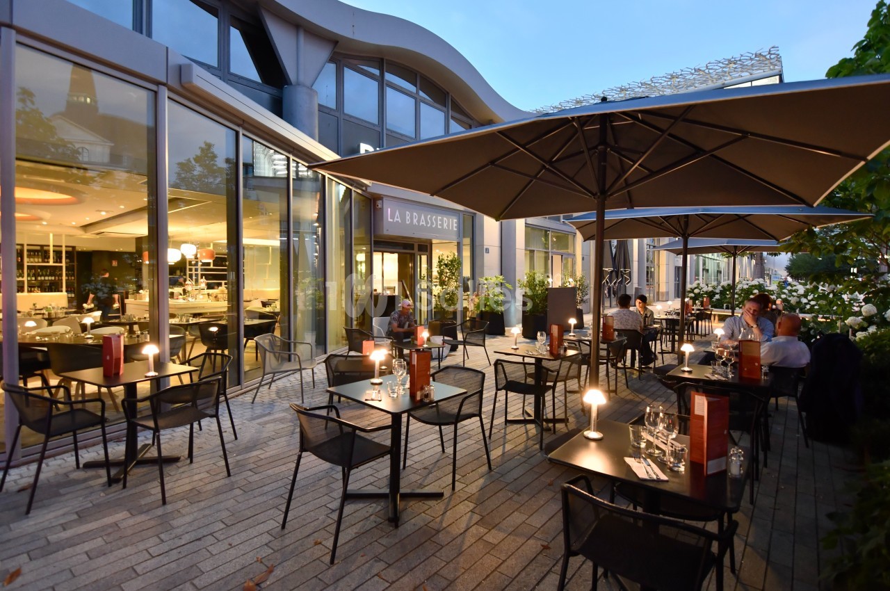 Terrasse d'une brasserie moderne avec tables éclairées, chaises noires et parasols, en soirée.