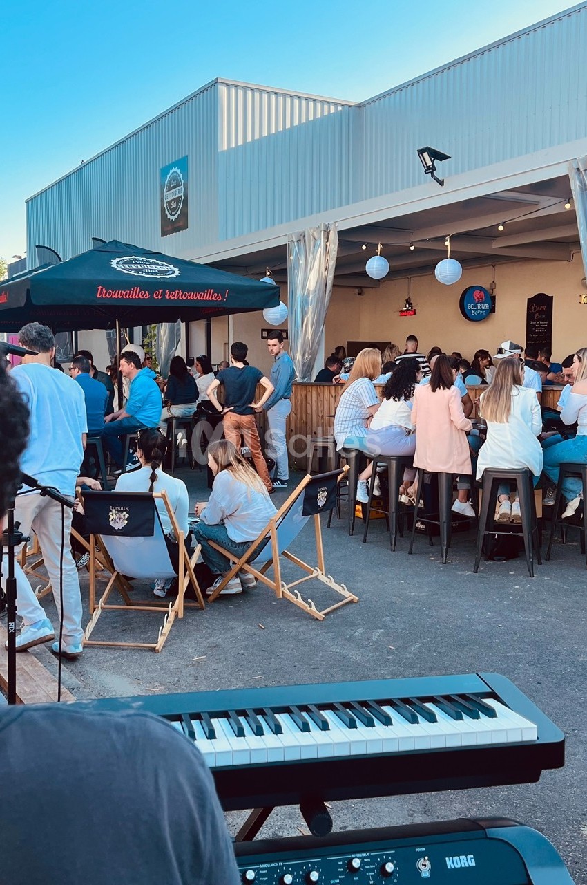 Groupe de personnes assises et debout dans une ambiance conviviale en extérieur près d'un bar, avec un clavier au premier…