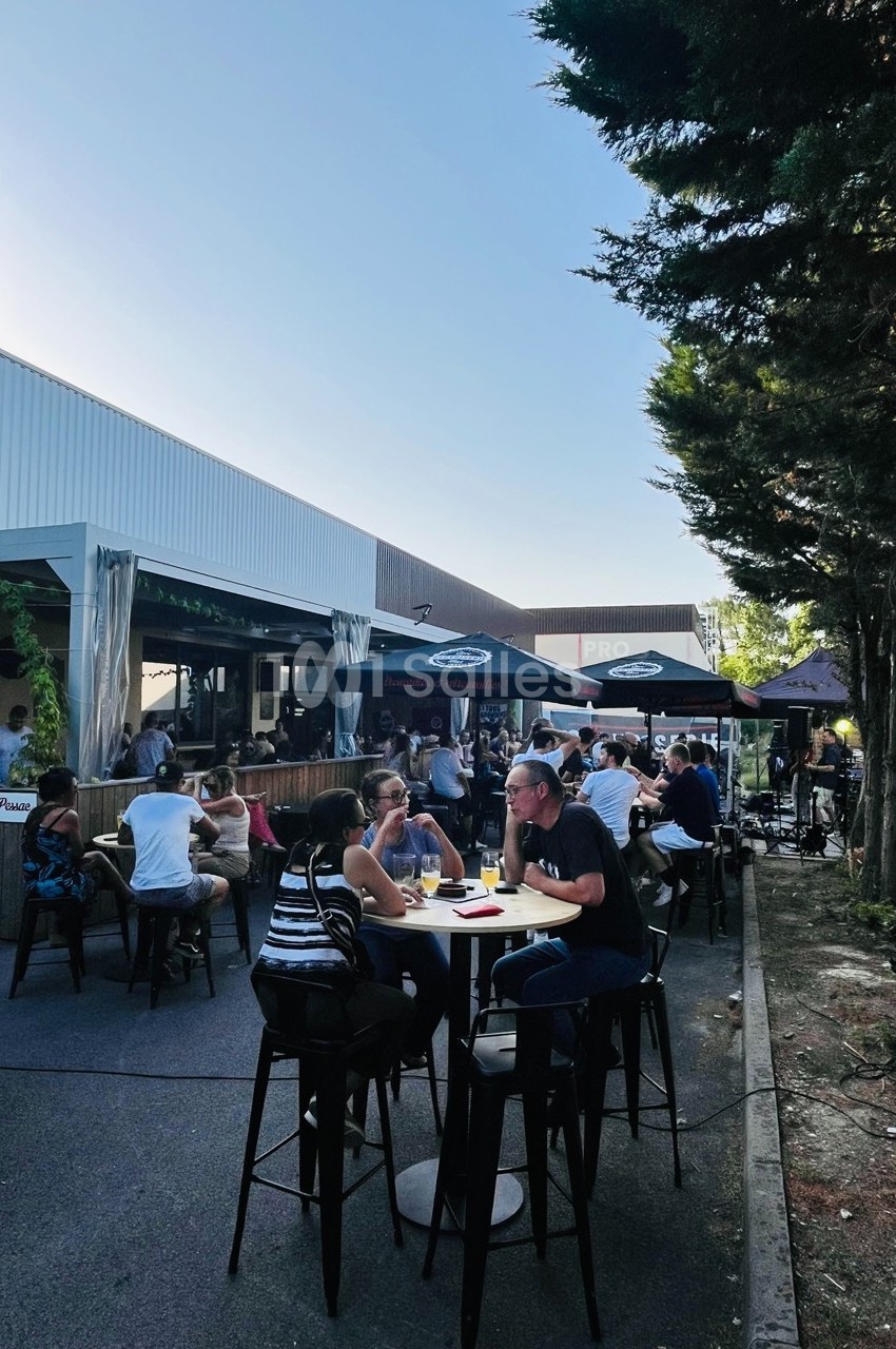 Personnes attablées en extérieur dans une ambiance conviviale, près d'un bâtiment moderne et d'arbres.