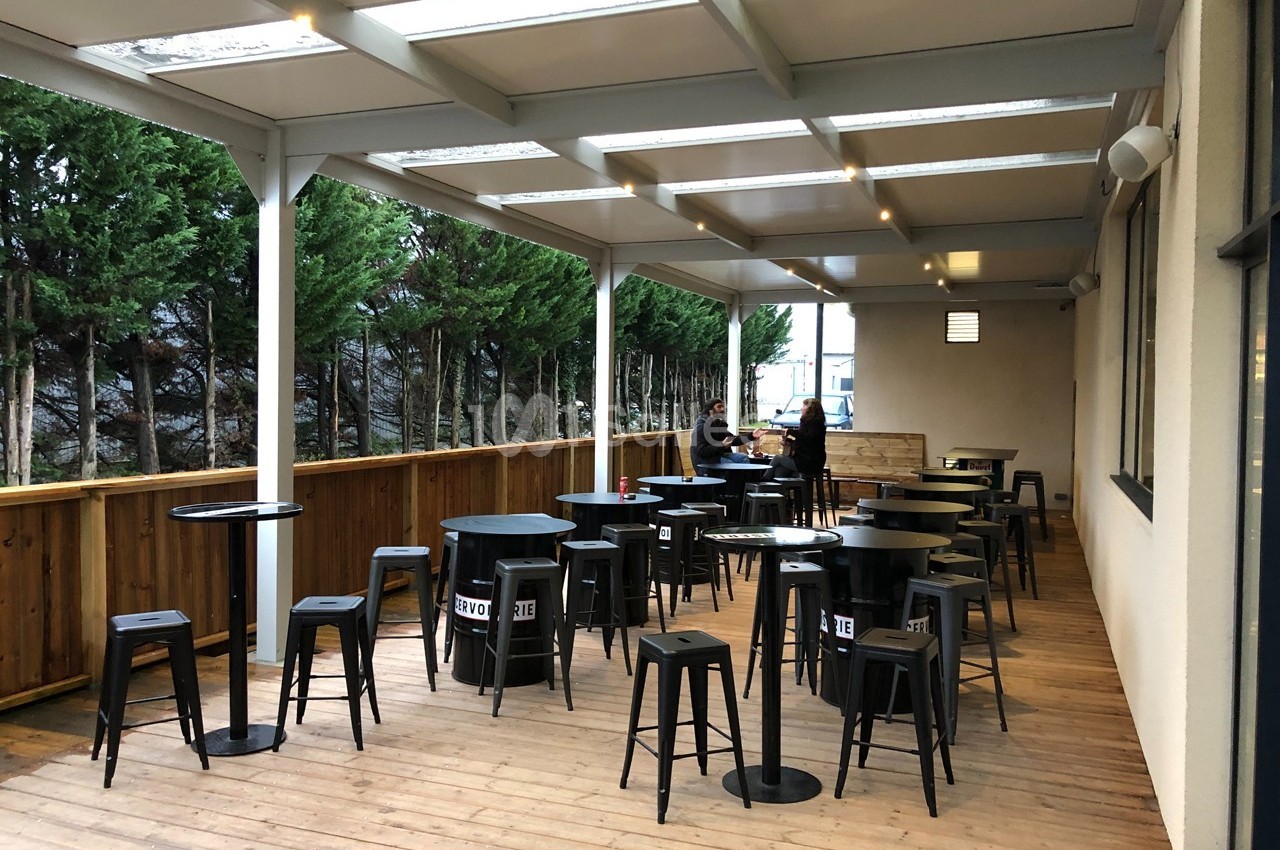Terrasse en bois couverte avec tables hautes, tabourets noirs et deux personnes discutant au fond.