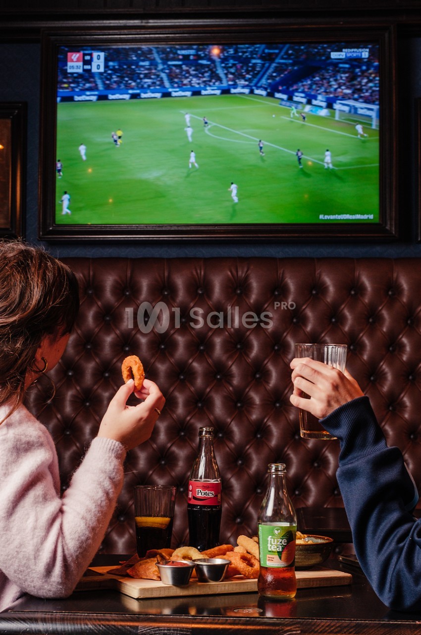 Deux personnes regardent un match de football à la télévision dans un bar, en mangeant et buvant à une table.