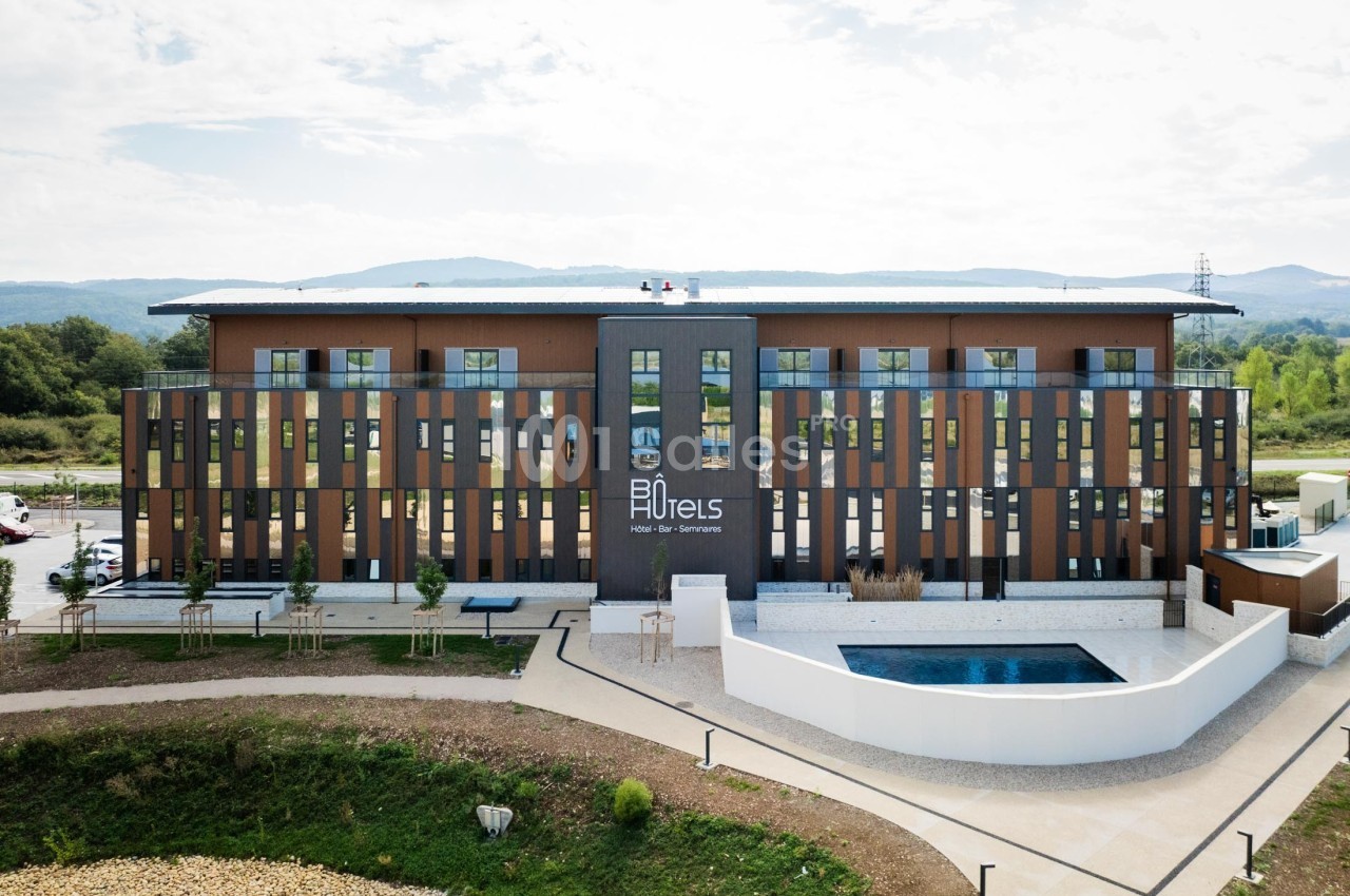 Façade d'un hôtel moderne à trois étages avec piscine extérieure, entouré de verdure et d'un ciel partiellement nuageux.