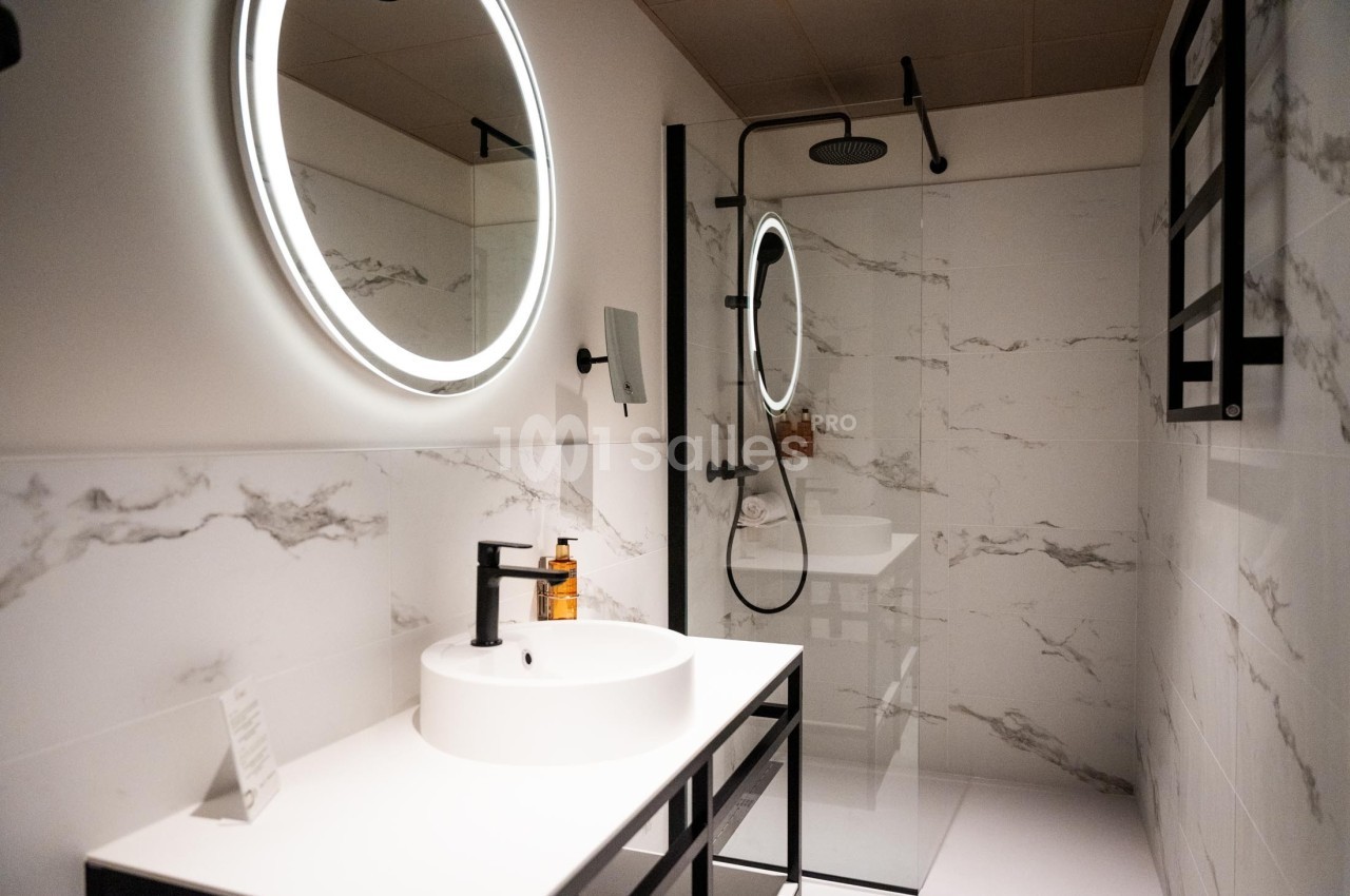 Salle de bain moderne avec lavabo blanc, miroir rond éclairé, douche à l'italienne et murs en carrelage effet marbre.