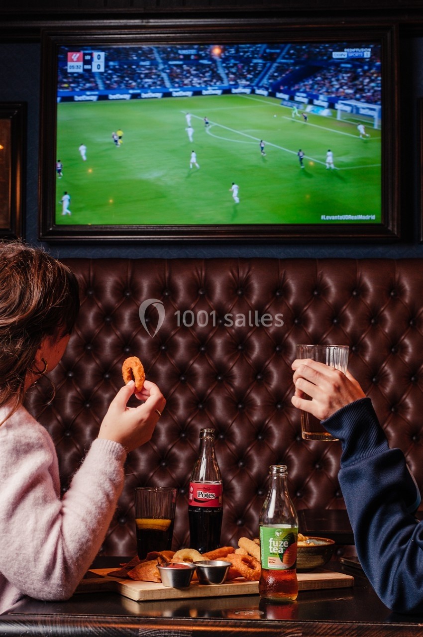 Deux personnes regardent un match de football à la télévision dans un bar tout en partageant des snacks et des boissons.