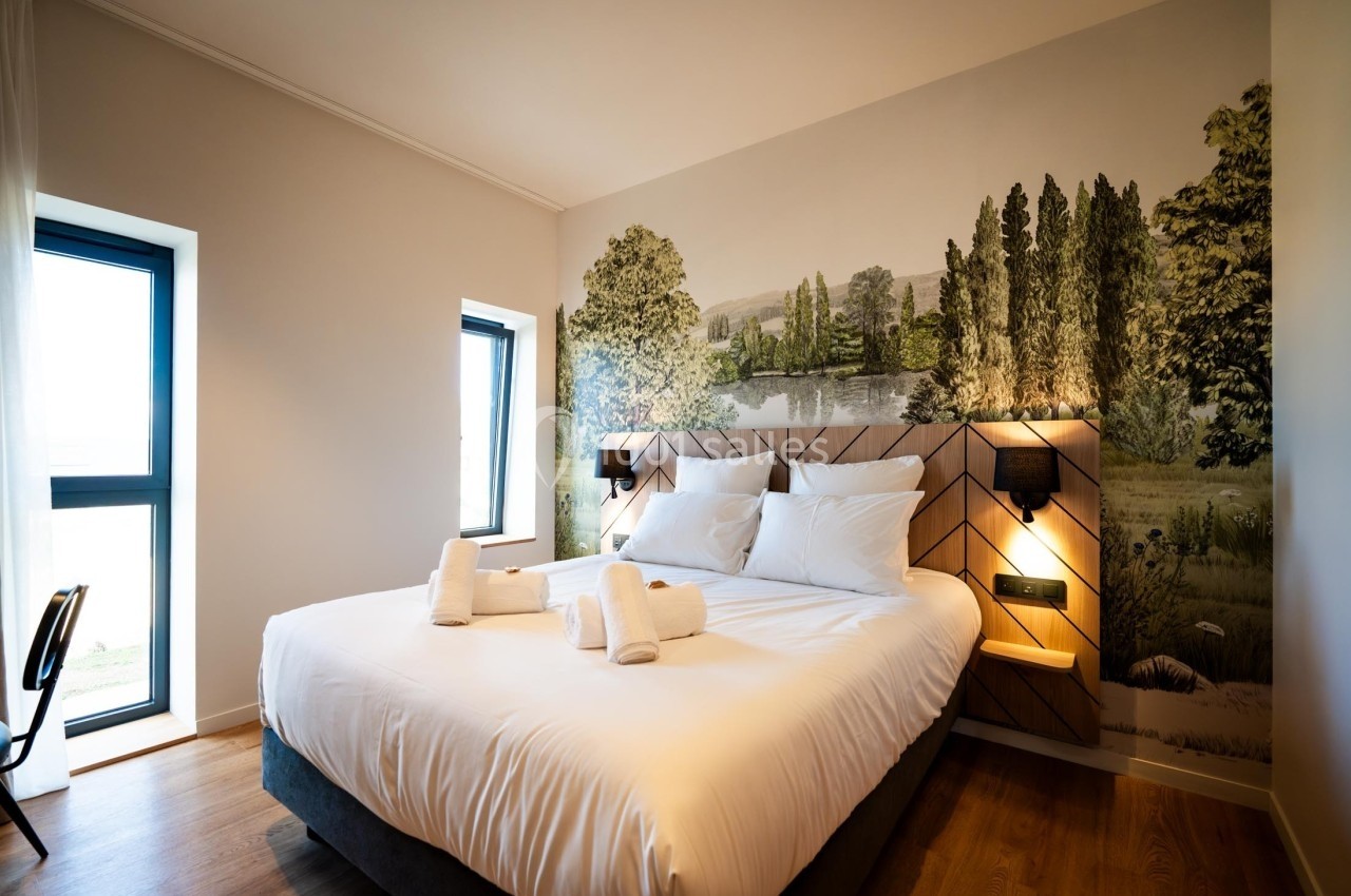 Lit double avec draps blancs, serviettes pliées, éclairage mural et mur décoré d'une fresque paysagère.