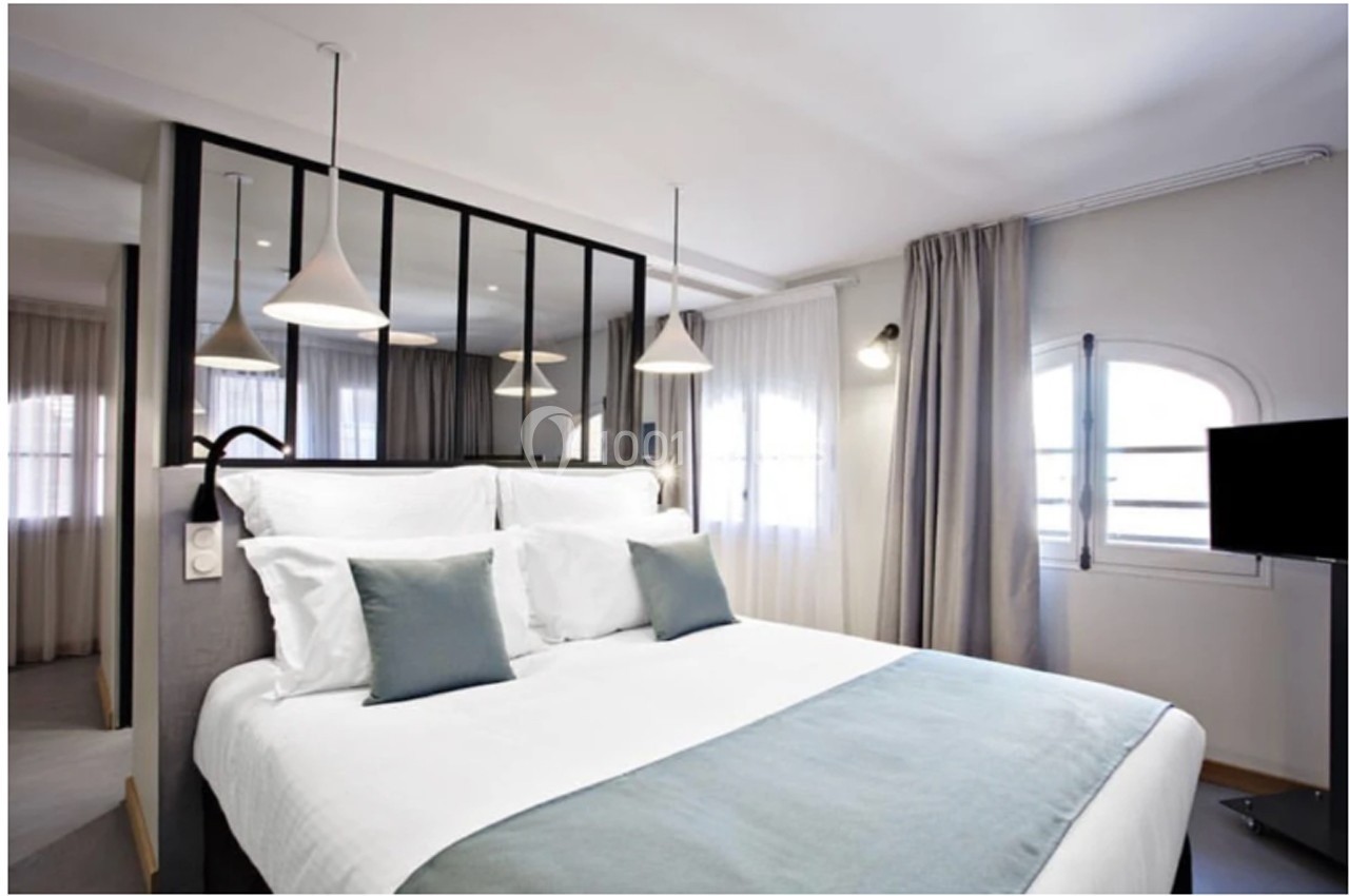 Chambre lumineuse avec lit double, coussins bleus, suspensions modernes et grande verrière séparative.