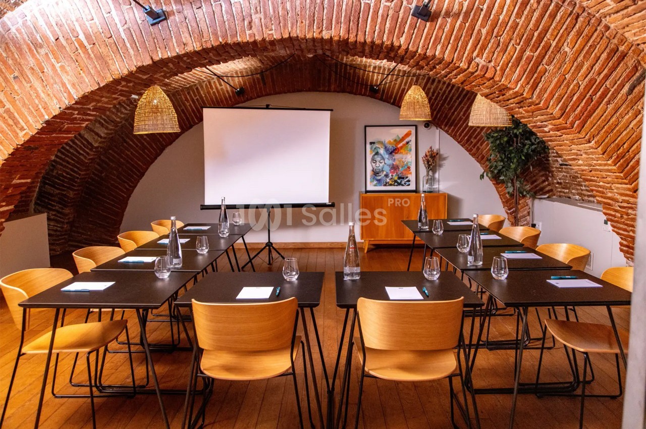 Salle de réunion voûtée avec tables, chaises en bois, écran de projection et décorations murales colorées.