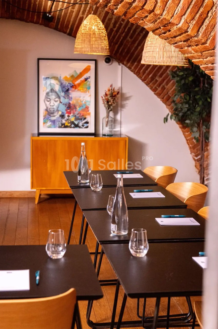 Salle de réunion avec tables noires, chaises en bois, bouteilles d'eau, carnets, et une décoration murale colorée.