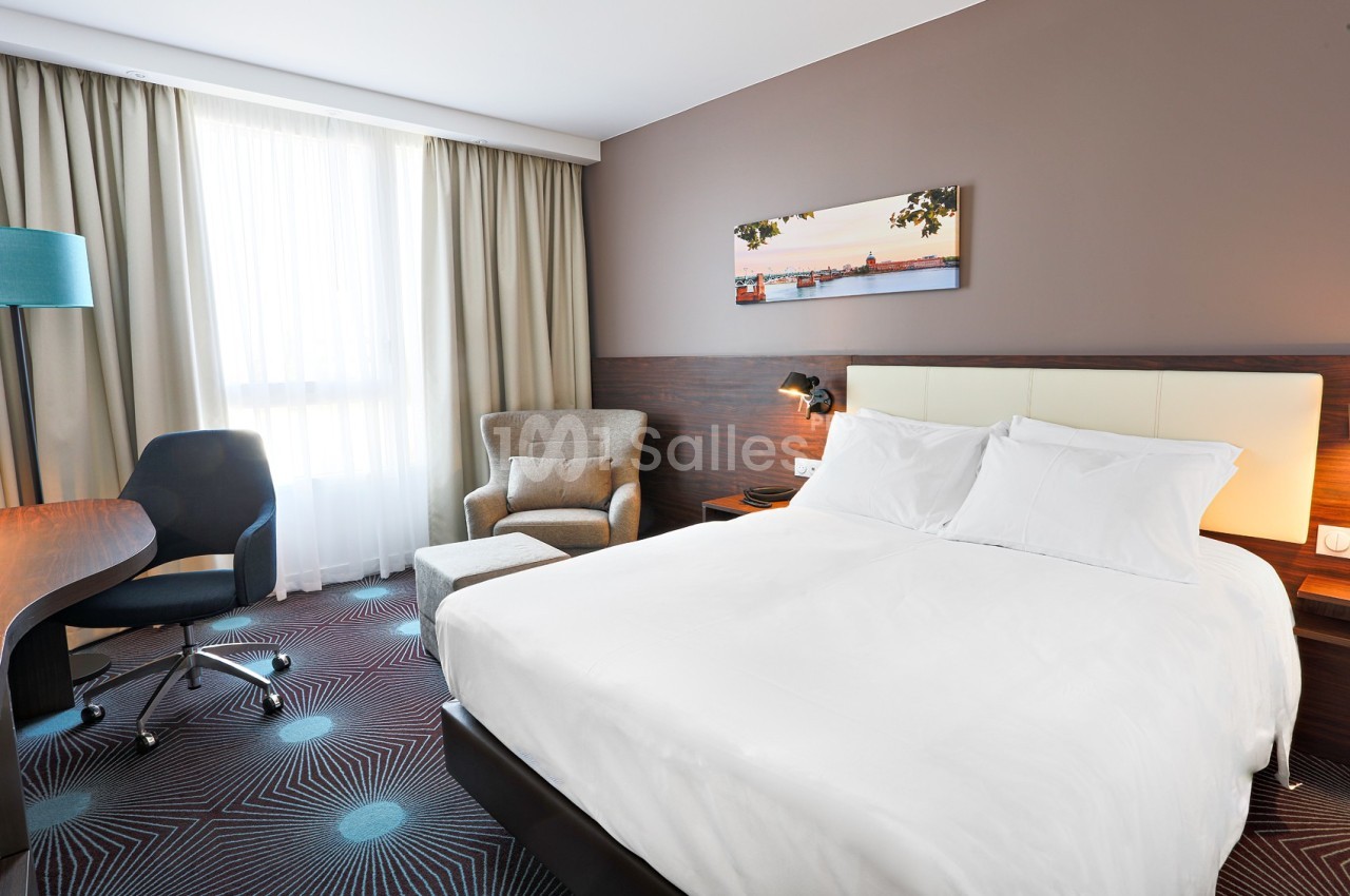 Chambre d'hôtel lumineuse avec un lit double, un fauteuil, un bureau, une chaise et une grande fenêtre avec rideaux.
