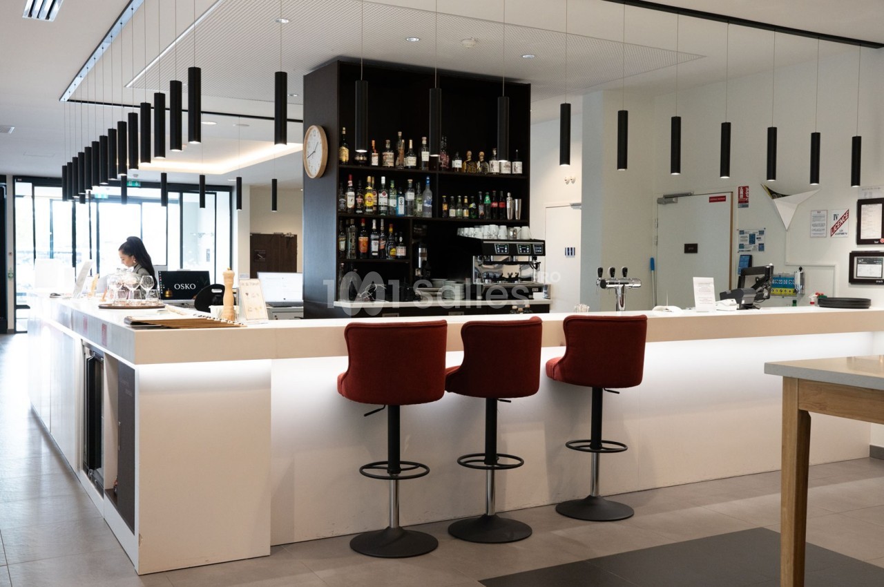 Bar moderne avec comptoir blanc, tabourets rouges, étagères de bouteilles et machine à café en arrière-plan.
