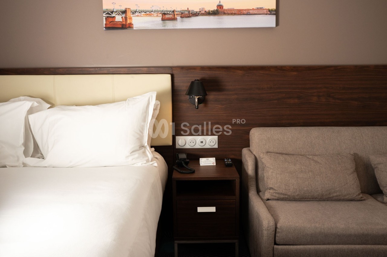 Lit avec draps blancs, table de chevet avec lampe et prises électriques, canapé beige dans une chambre moderne.